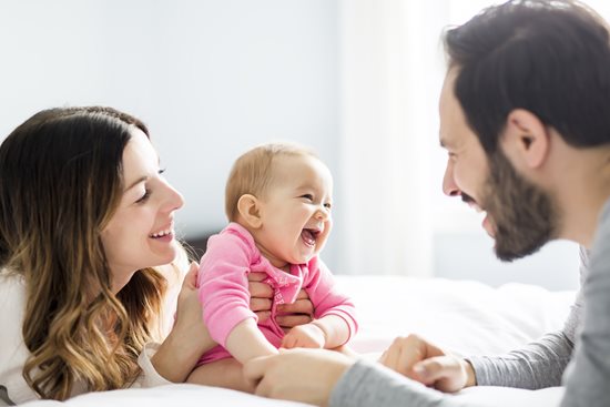 A smiling baby being held by her mother on a bed, while looking at her father