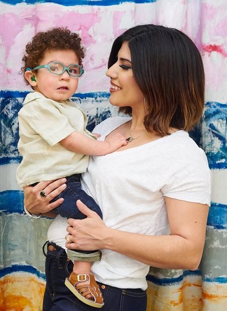 Una mujer sonriente sosteniendo a su hijo pequeño con anteojos frente a un mural colorido,