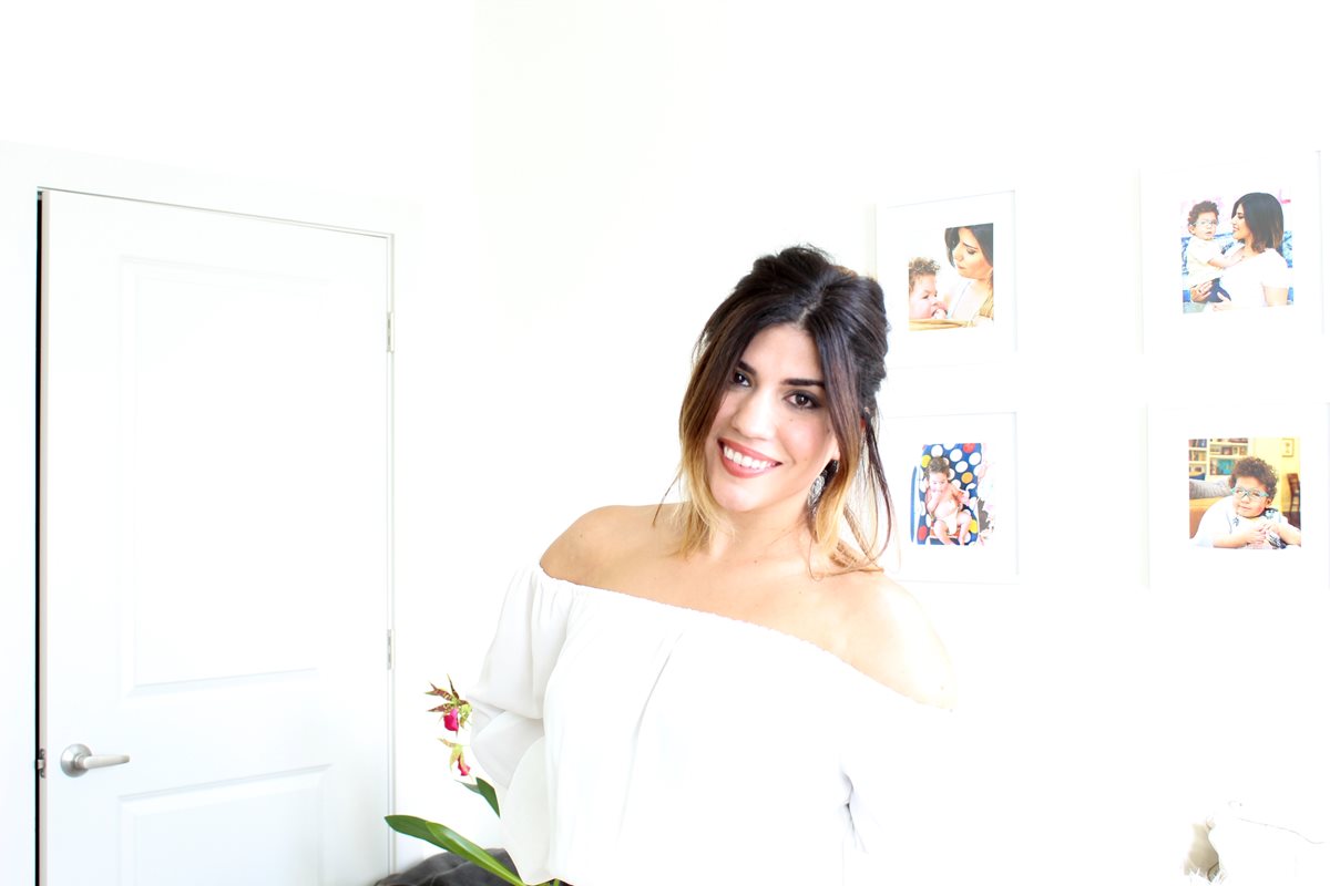 A women in a white dress smiling, in front of a white wall in a living room 