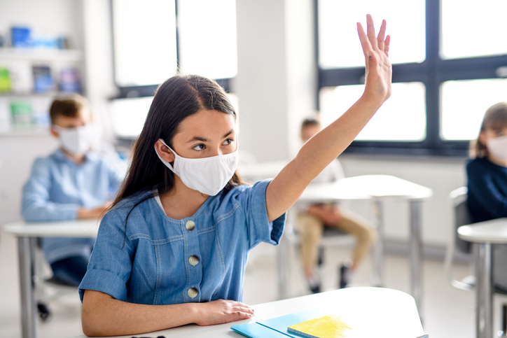 A masked student in a classroom raising their hand 