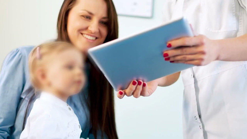 enfermera le muestra a una madre con una hija pequeña la pantalla de una tableta