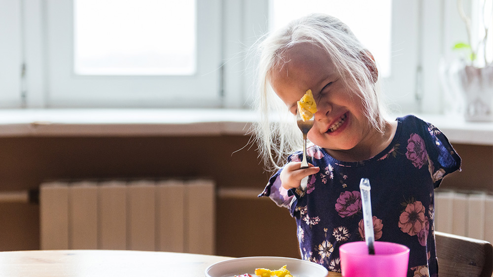 onfident little girl with no arms and two fingers on her hand proudly holds a fork while eating breakfast.