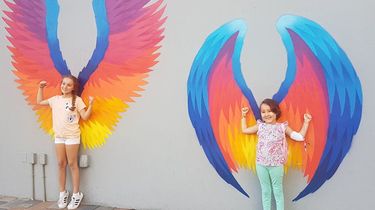 former patient Isabella, and her sister, posing against a wall with multicolor angel wings