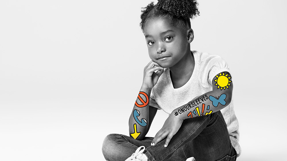 black & white photograph of african american girl sitting cross legged, colorful symbols #onoursleeves mark her arms.