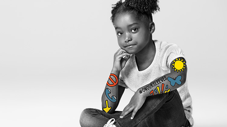 black & white photograph of african american girl sitting cross legged, colorful symbols #onoursleeves mark her arms.