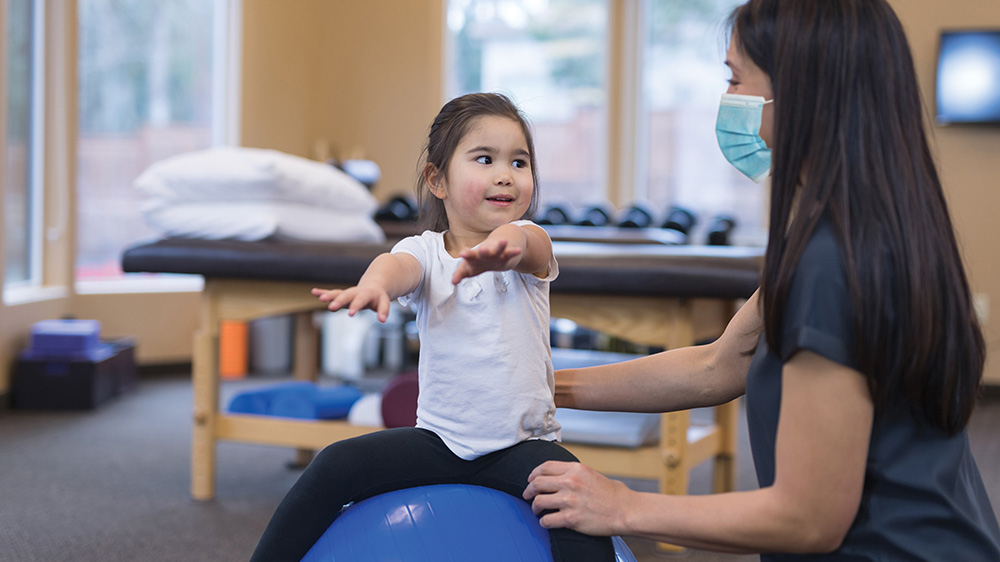 little girl sitting on exercise ball during rehab exercises with therapist