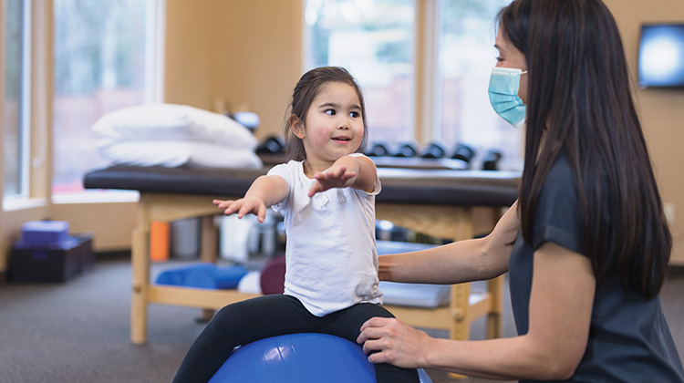 niña pequeña sentada sobre una pelota de ejercicio durante ejercicios de rehabilitación con la terapeuta