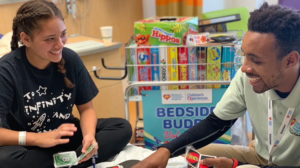 teen volunteer playing a card game with a patient at their bedside.