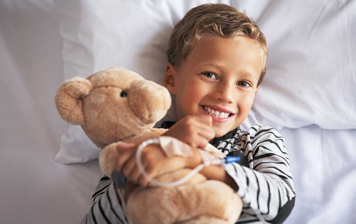 Boy smiling with an IV holding a bear in a hospital bed