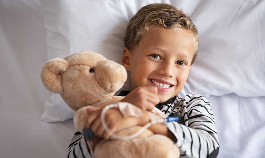 Boy smiling with an IV holding a bear in a hospital bed