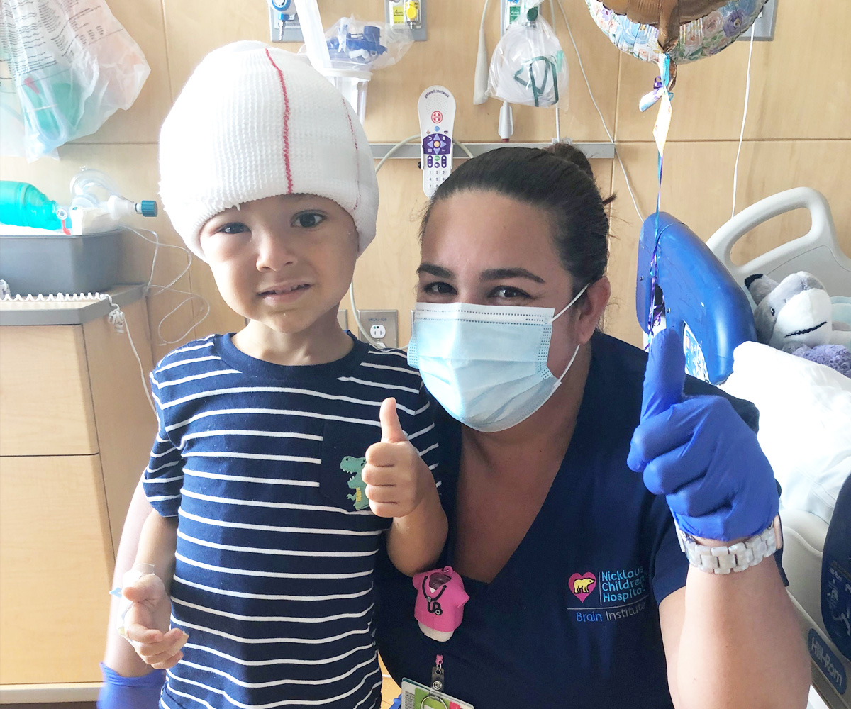 Jayden with his head bandaged giving a thumbs up next to his nurse.