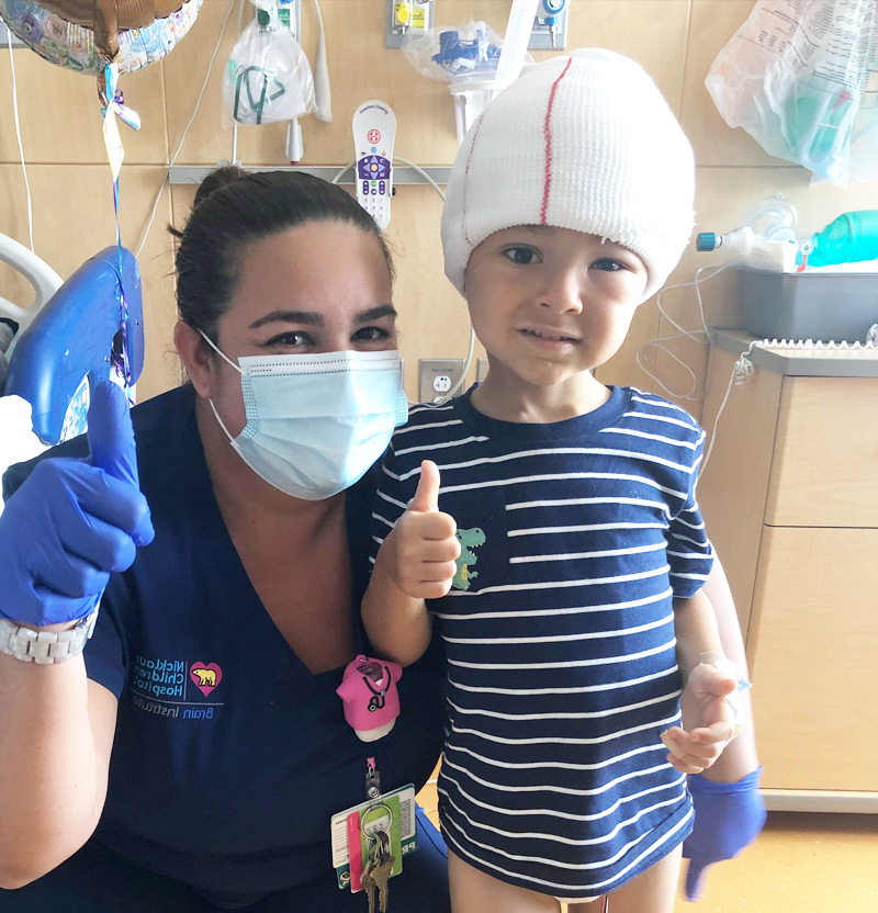Jayden with his head bandaged giving a thumbs up next to his nurse.