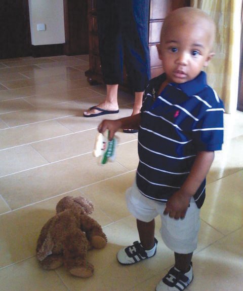 Carmani as a toddler standing up on a tile floor looking at the camera while he plays.