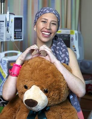 Raquel in a hospital room holding a large teddy bear and making a heart sign with both of her hands