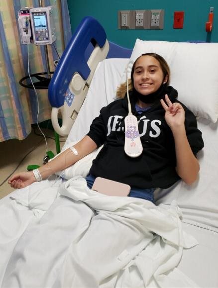 Nyla in a hospital bed smiling and giving the peace sign.