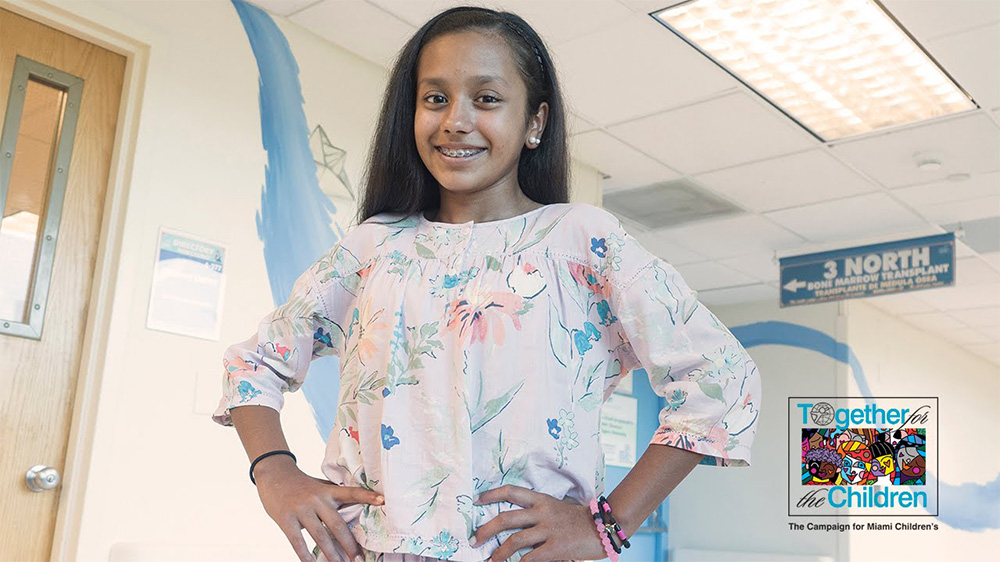 Alexandra wearing a flower patterned blouse with both her hands on her waist while she smiles.