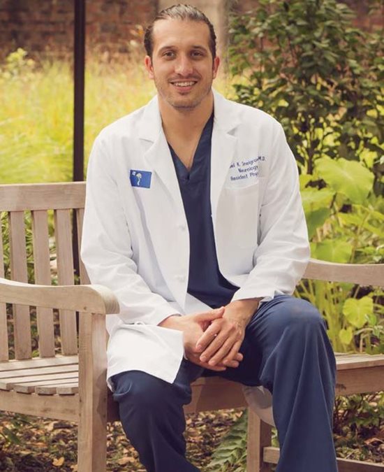 Danuel Snelgrove sitting on a park bench wearing medical scrubs and a lab coat. 