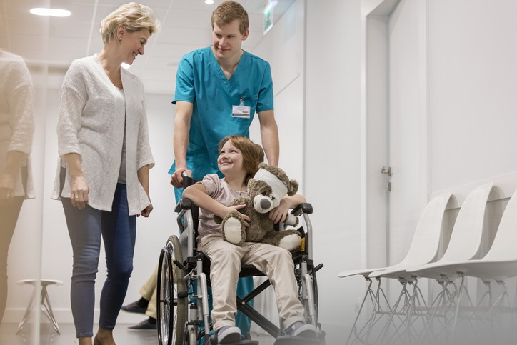 Child leaving examination room in a wheelchair pushed by attendant, smiling at mother.